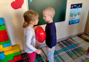 Dziewczynka z chłopcem tańczą w parze, trzymając serduszkowy balon między brzuchami.